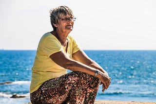 Older woman with glasses sitting on a beach with ocean behind her.