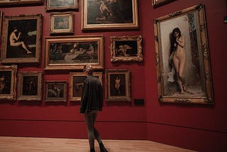 A young woman in an art gallery, admiring nineteenth century paintings. She has blonde hair worn up in a fishtail French braid, and she is wearing black ankle boots, grey leggings, and a blue denim jacket which is a little too large for her, so the sleeves cover her hands.