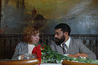A man and a woman sit at a table with a bouquet of flowers before them.