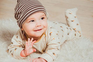 A smiling baby lying on a soft white fur rug. The infant is wearing a brown and white striped beanie hat and a light beige onesie with small brown and orange patterns. The baby is looking up at the camera with a happy expression and has one hand raised as if waving. The setting appears to be a home with wooden flooring visible in the background.