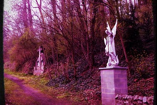 Photo with lavender filters of a bleak winter path with a statue of a cross and another of an angel. Photo was taken by Martina Clark in Belgium.