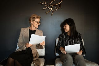 Two women sitting on chairs looking at papers. The left one is older, wearing a business suit, and looks more professional, but she’s still listening attentively to the younger one