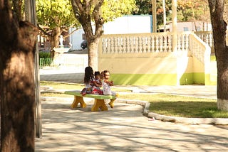 Foto de uma praça com três crianças sentadas em um banco na frente de um coreto.