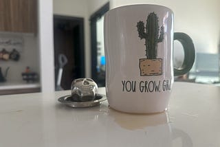 A mug with a cactus painted on it reading, “You grow, girl”. Tea is spilled all over the counter under it.
