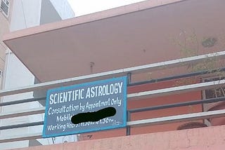 An image shows a hand-lettered sign with a blue background and white text mounted on a railing of a building. The building has a light brown exterior and a pink overhang. The sign displays the following text: “SCIENTIFIC ASTROLOGY”, “Consultation by Appointment Only”, “Mobile” (with a blacked-out section), and “Working Hours: 11.30 AM to 1.30 PM”. In the background, parts of other buildings and some greenery are visible.