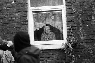 A monochrome picture. An elderly woman with glasses looks out of the window of a stone house. Next to the window hangs a plant pot in the shape of a cornucopia. Vines grow out of it. A group of people dressed like youngsters is passing by the window, their faces can’t be seen.