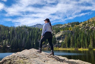 Me standing on a rock overlooking a lake with alpine trees and mountain tops in the distance