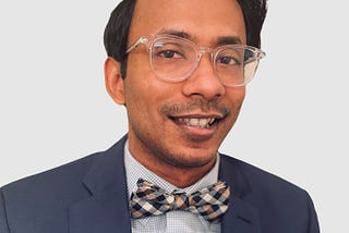 Headshot of Ather, who is Asian, smiling, wearing a navy suit and checkered bowtie. He is wearing thick clear-framed glasses.