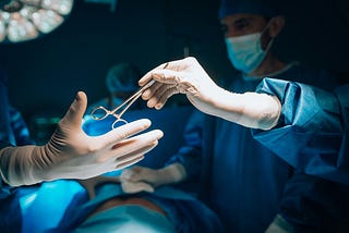 A healthcare professional in blue scrubs passes scissors to another during surgery.