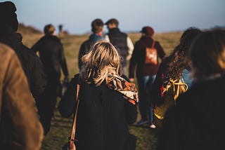 Photo of a group of people, in coats and scarves, walking up a field. The focus is soft around the edges, and only the person in the center is clear — black coat, orange patterned scarf, tousled blond hair around their shoulders.