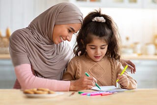A woman wearing a Hijab helping a schoolchild (possibly with autism or Asperger’s) with her work.