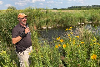 a man with a microphone stands at the edge of a pond surrounded by tall flowers and grasses