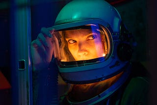 Close-up photo of woman wearing a spacesuit helmet in the dark. The inside of the helmet has an internal orange glow, illuminating her face. She is holding the top edge of the helmet window with her right hand and she is looking to her left.