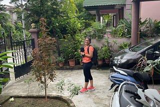 A woman in gym gear in her driveway stops to augh at the photographer on her way to the gym. There is a car and a scooter in the driveway. There are green planst and trees in the garden. Its summer in the picture.