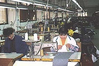 Two rows of industrial sewing machines extending backwards in a factory in China. The first two machines are being operated by a man and a woman who are smiling as they work.