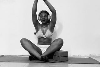 A black andd white picture of Don Precious seated legs crossed on a yoga mat with yoga blocks holding up her left knee