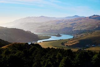 Long distance vista of golden mountain ridges below blue sky, towering over a blue winding sinuous river, through a distant valley with a small village, surrounded by forest at dusk.