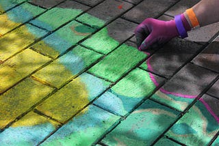 Close up image of a hand tracing a chalk outline around a colourful pattern on a brick pavement.