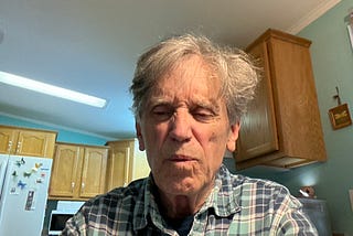 A man with gray hair is sitting in a kitchen, wearing a green and blue plaid shirt. He seems to be slightly grimacing, as if reacting to a pain or something unpleasant. The background includes wooden kitchen cabinets, a refrigerator with butterfly magnets, and a microwave.