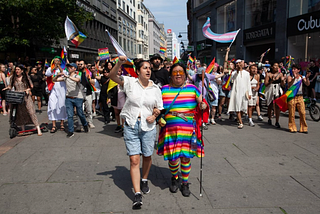 Group of people marching with various rainbow and Trans pride flags. In front: General Secretary from Salam, an organisation serving queer Muslims. Begard Reza (left) marches beside the leader of Steatornis, Maria Puenchir. Credit: Shahram Kiani.