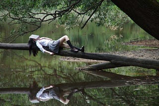 woman, with an open book covering her face, lying on tree trunk that leans out over a reflective lake