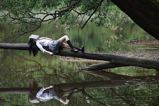 woman, with an open book covering her face, lying on tree trunk that leans out over a reflective lake