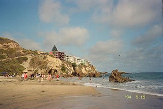 The sand, water, and cliffs of Corona del Mar. Large houses sit at the top of the cliffs, and large white clouds fill the blue sky.