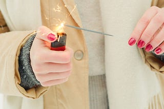 A woman with red polished nails holds  a sparkler in one hand and lights it with a red lighter in the other.