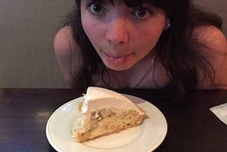 Anna leans head over banana cream pie on plate, sticking tongue out and making wide googly eyes at it.