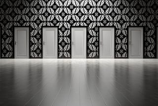 Black and white photo of a wall with decorative wallpaper. On the wall are five white doors. In the foreground of the photo are bare wood floors.