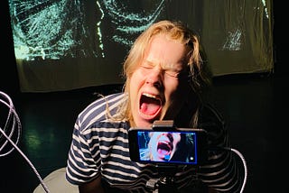 A female dancer crouches on the floor showing  a silent scream. Her open mouth is visible on a video camera and in turn the image is projected behind her on to a screen.