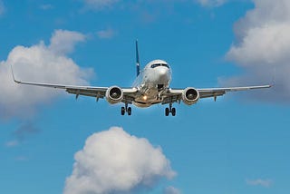 A large passenger plane is flying toward the camera. Wheels are down as it prepares to land. There are a few puffy white clouds in the blue sky.