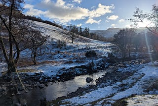 A shallow stream flows through a snowy hillside landscape with bare trees and distant hills under a blue sky.