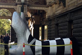 Giant feet, wearing silver shoes and striped socks, emerging from a building.