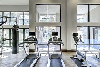 A bright and spacious gym featuring two treadmills, a stationary bike, and a punching bag, with large windows allowing natural light to fill the space