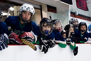 Hockey players lean against the boards to watch a game in progress. They are all wearing the same blue, white and green jerseys.