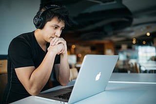 Image of a young man wearing black wireless headphones and is deeply focused on his macbook that is in front of him.