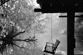 A porch swing hangs from the roof of a ramshackle dock on a river. In the distance are trees whose branches extend out over the river. The air appears foggy or misty. The photo is in black and white.