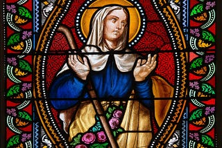 A stained glass window. It depicts a haloed woman holding her hands up to heaven with an exasperated expression. Her hands are about fourteen inches apart.