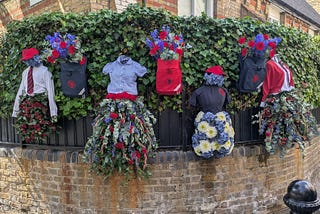 A rounded corner brick wall with a black fence on top covered by ivy on which are hanging school uniforms and school satchels decorated with flowers.