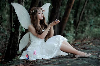A beautiful dark-haired woman dressed in a white fairy costume sits in the forest holding colorful butterflies