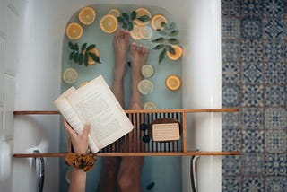 knees down person in tub with book wrack over the top and book. Lemons and greenery in bath water.