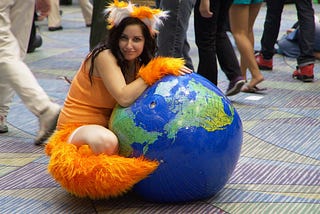 A woman in an orange-eared and cuffed Firefox cosplay hugging an inflatable globe, smiling as she sits on a patterned floor.