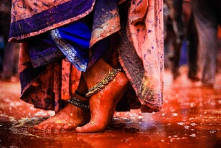 A cropped image of a woman’s feet standing in a shallow puddle with wide silver anklets and an overhanging sari.