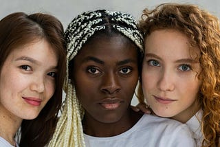 A portrait of three young women (an asian woman, a black woman, and a white woman) with subtle expressions, resting their heads against one another and looking into the camera.