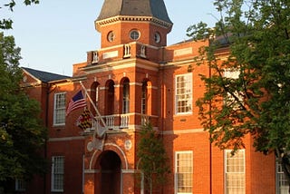 A historic brick buiding with trees on either side of the photo and white-trimmed windows. An American flag and a Maryland flag can be seen on the front of the building.