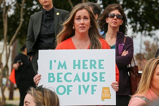 Picture of a woman looking at the camera holding a sign that reads “I’M HERE BECAUSE OF IVF”