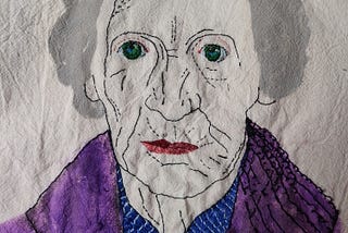 Embroidered portrait of an elderly white woman with a blue shirt and purple coat