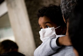 Small child with dark skin and curly black hair wears a face mask while being carried by an adult. The child looks over the adult’s shoulder, clinging to the adult, eyes wide.