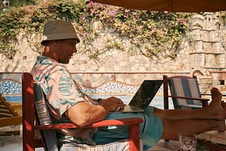 A man wearing an Hawaiian shirt sitting poolside with a compter on his lap and a drink by his side.